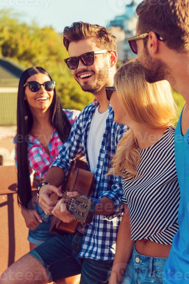 Enjoying time together. Side view of four young people bonding to each other and looking at camera while standing on the roofwith guitar photo