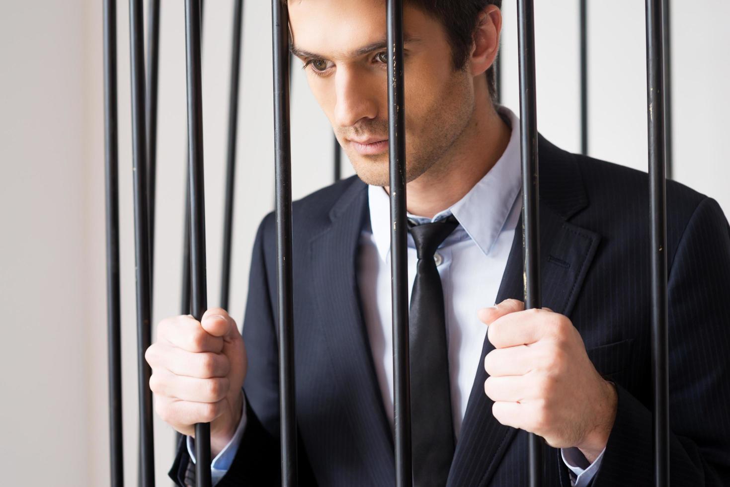 Business criminal. Depressed young man in formalwear standing behind a prison cell and looking at away photo
