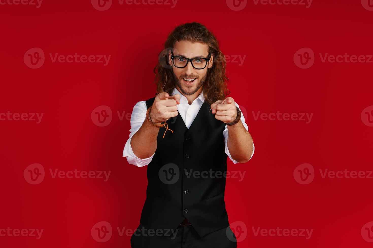 Cheerful young man in elegant clothing looking at camera and pointing you while standing against red background photo