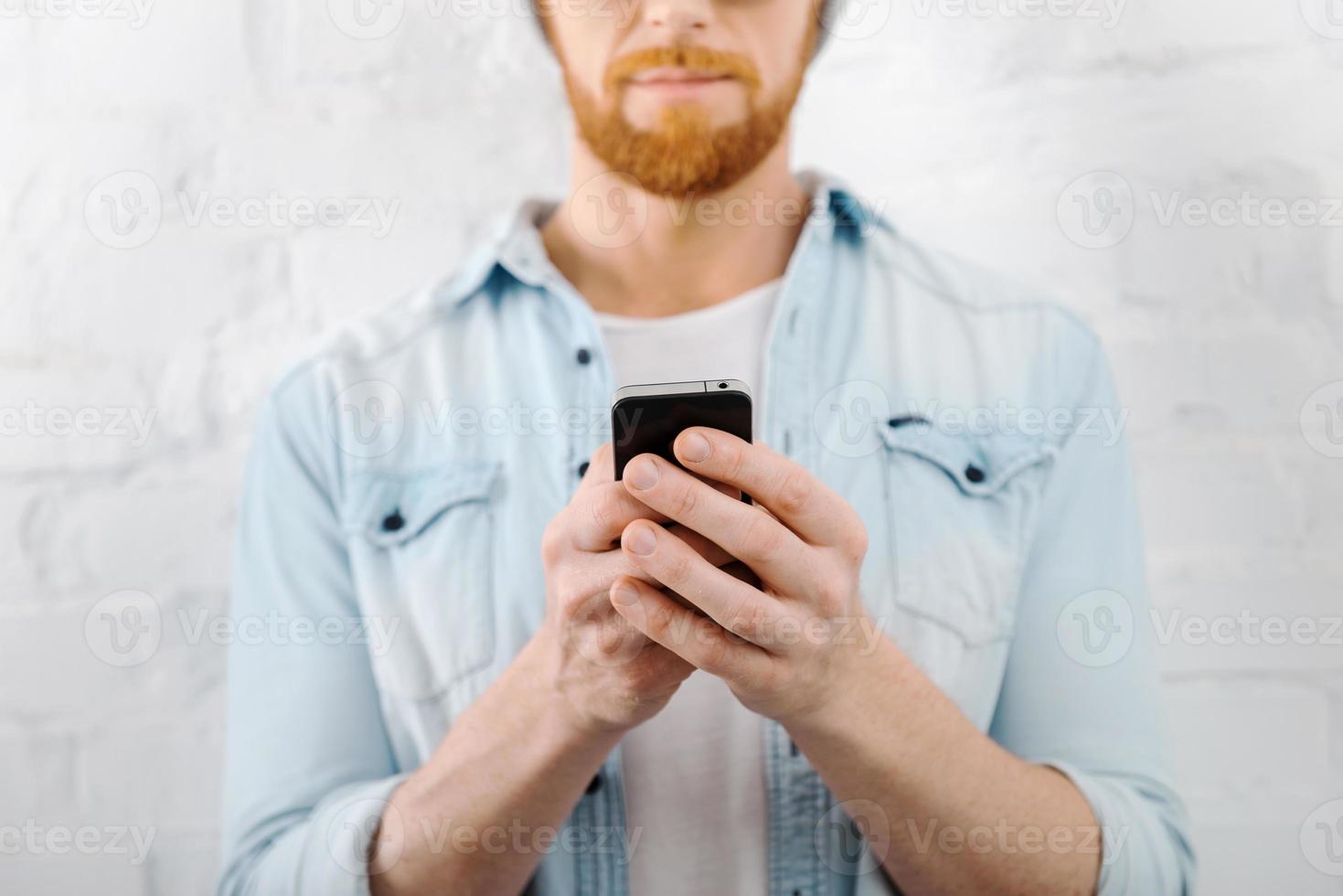 Man texting. Cropped image of bearded man holding mobile phone while standing against brick wall photo
