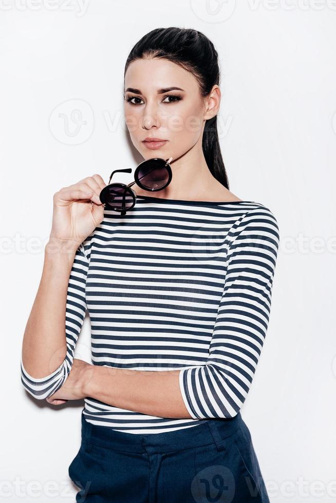 Keeping it cool. Beautiful young woman taking off her sunglasses and looking at camera while standing against white background photo