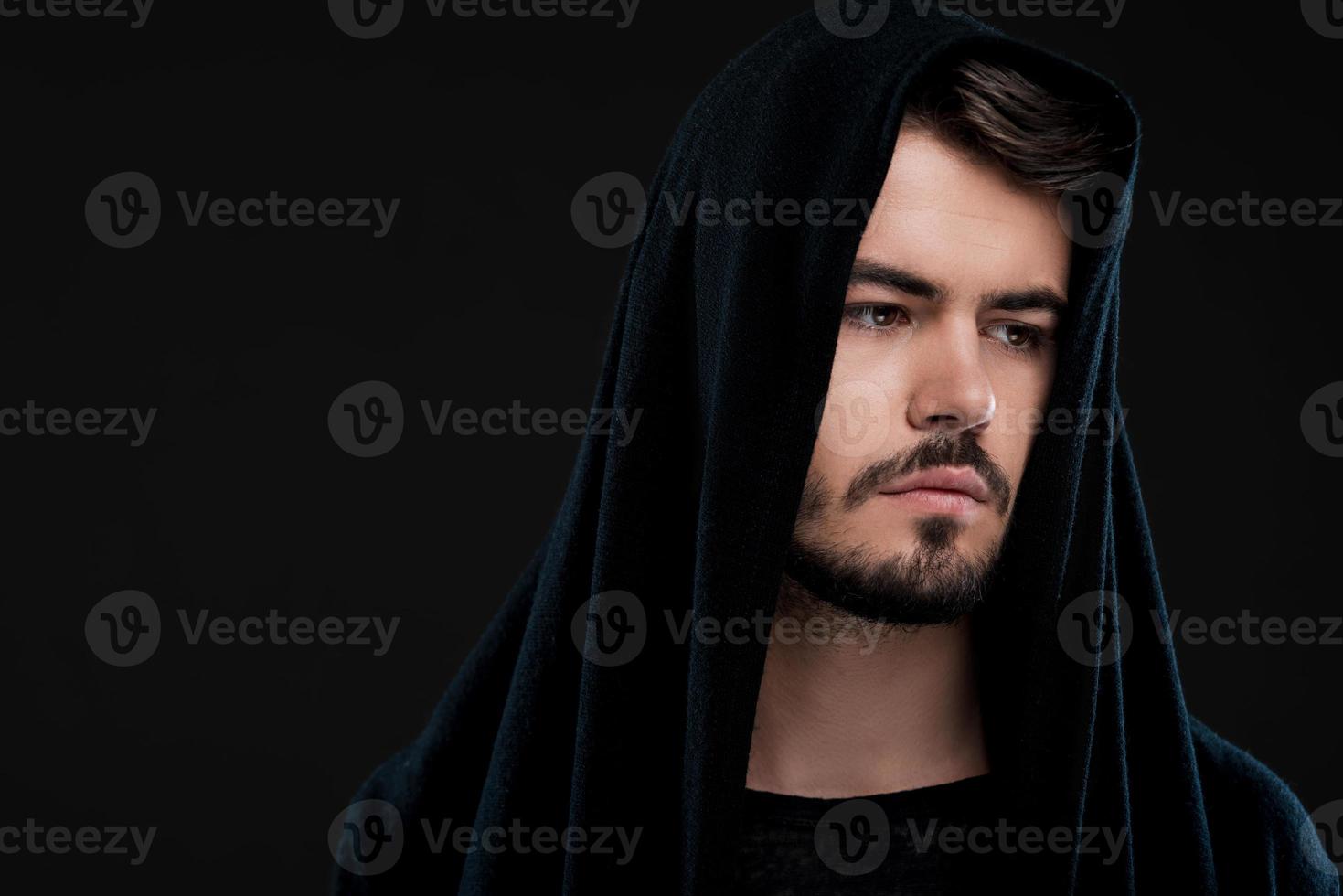 Handsome man in black. Pensive young man in hood looking away while standing against black background photo