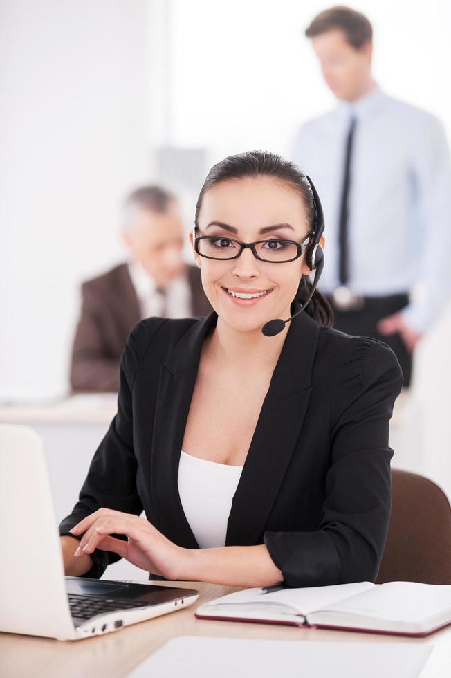 Customer service representative at work. Attractive young woman in formalwear adjusting her headset and smiling while people working on background photo