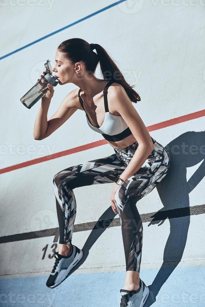 Tired after training. Top view of young woman in sports clothing drinking water while sitting on the running track outdoors photo