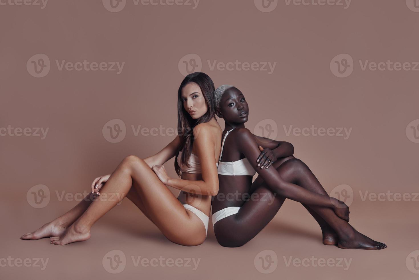 True feminine beauty. Two attractive mixed race women looking at camera while sitting back to back against brown background photo