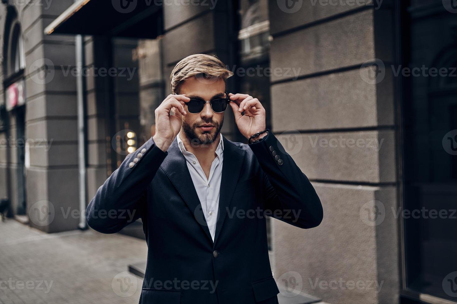 His style is perfect. Handsome young man in smart casual wear adjusting his eyewear while walking through the city street photo