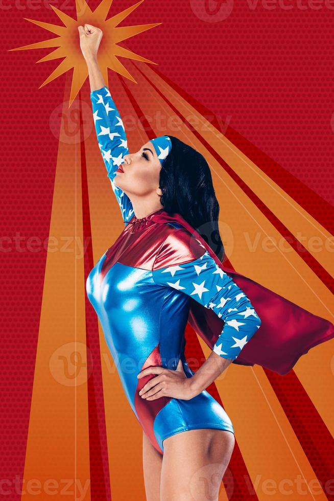 Going to save the world. Side view of beautiful young woman in superhero costume keeping arm outstretched while flying up against red background photo