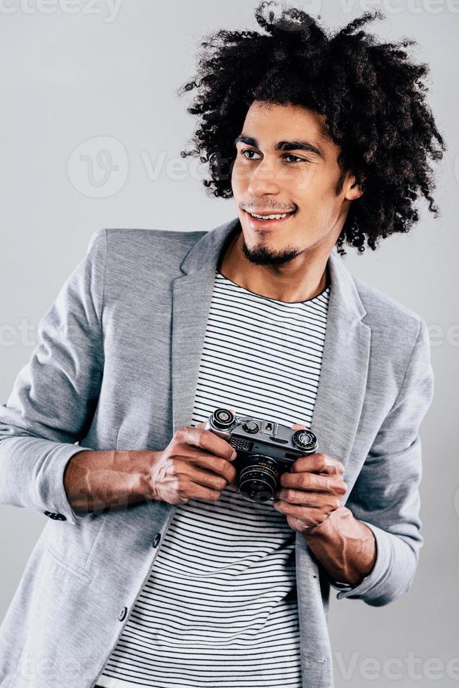 Cheerful photographer. Handsome young African man holding retro styled camera and looking away with smile while standing against grey background photo