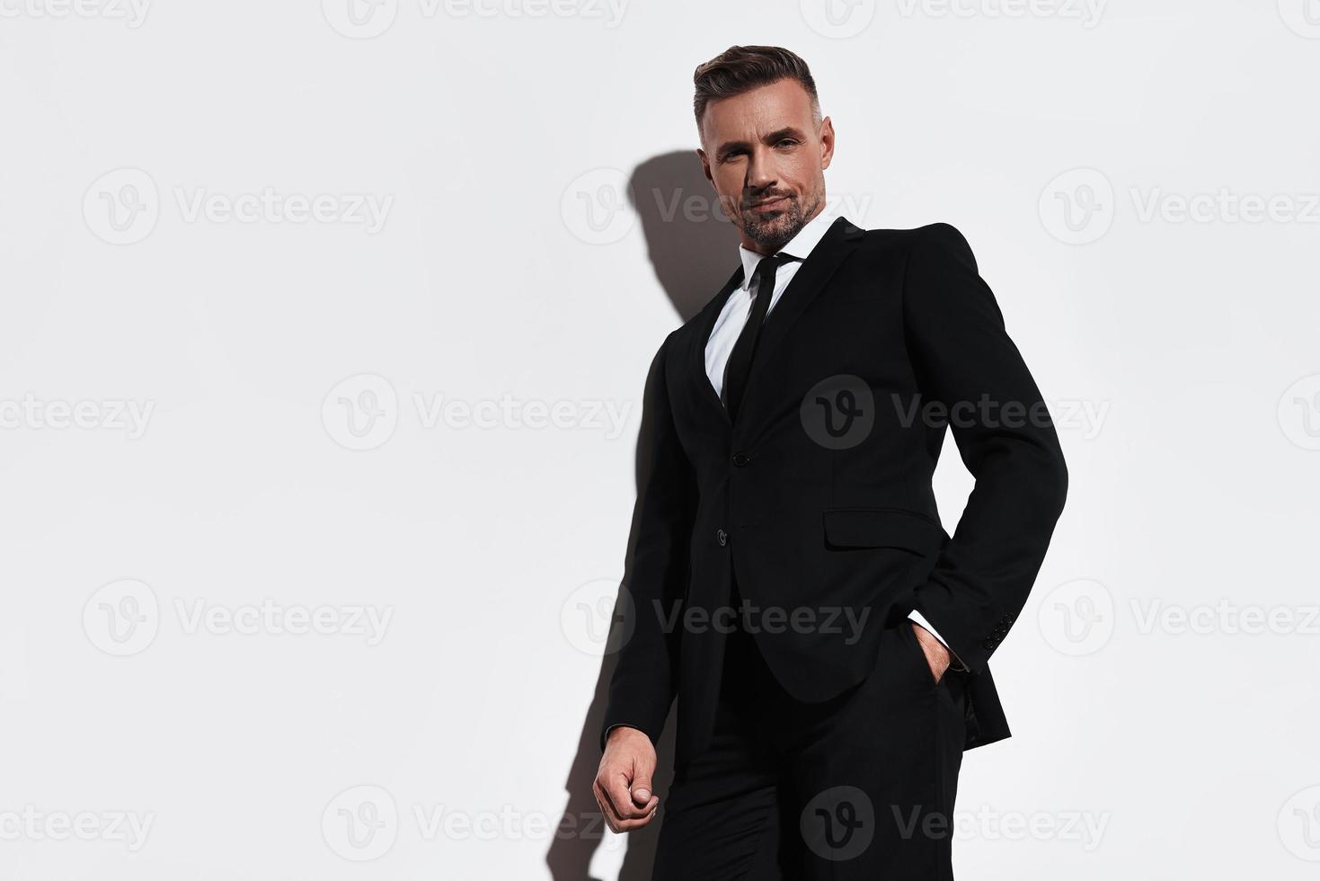 Used to look perfect. Handsome young man in full suit keeping hand in pocket and looking at camera with smile while standing against white background photo