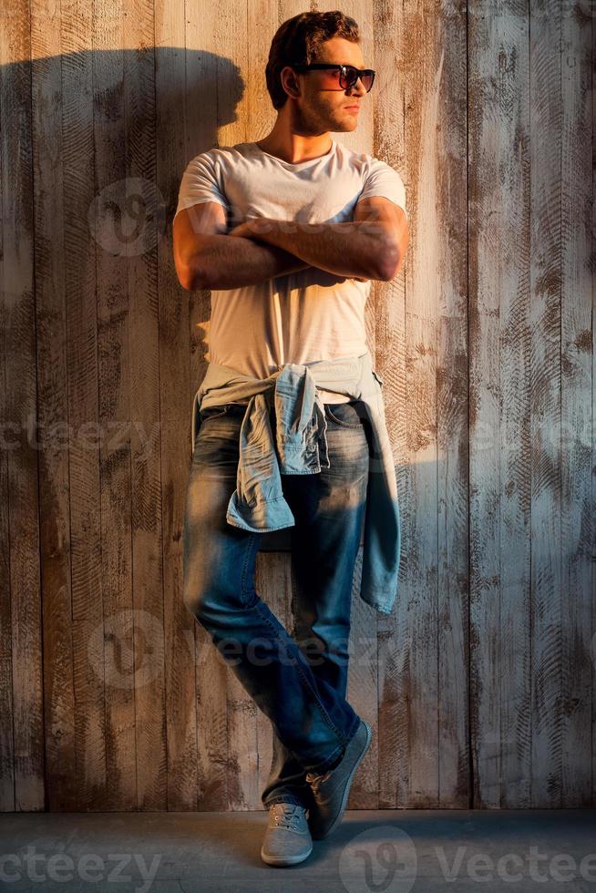 Casually handsome. Full length of handsome young man in sunglasses keeping arms crossed while standing against the wooden wall photo