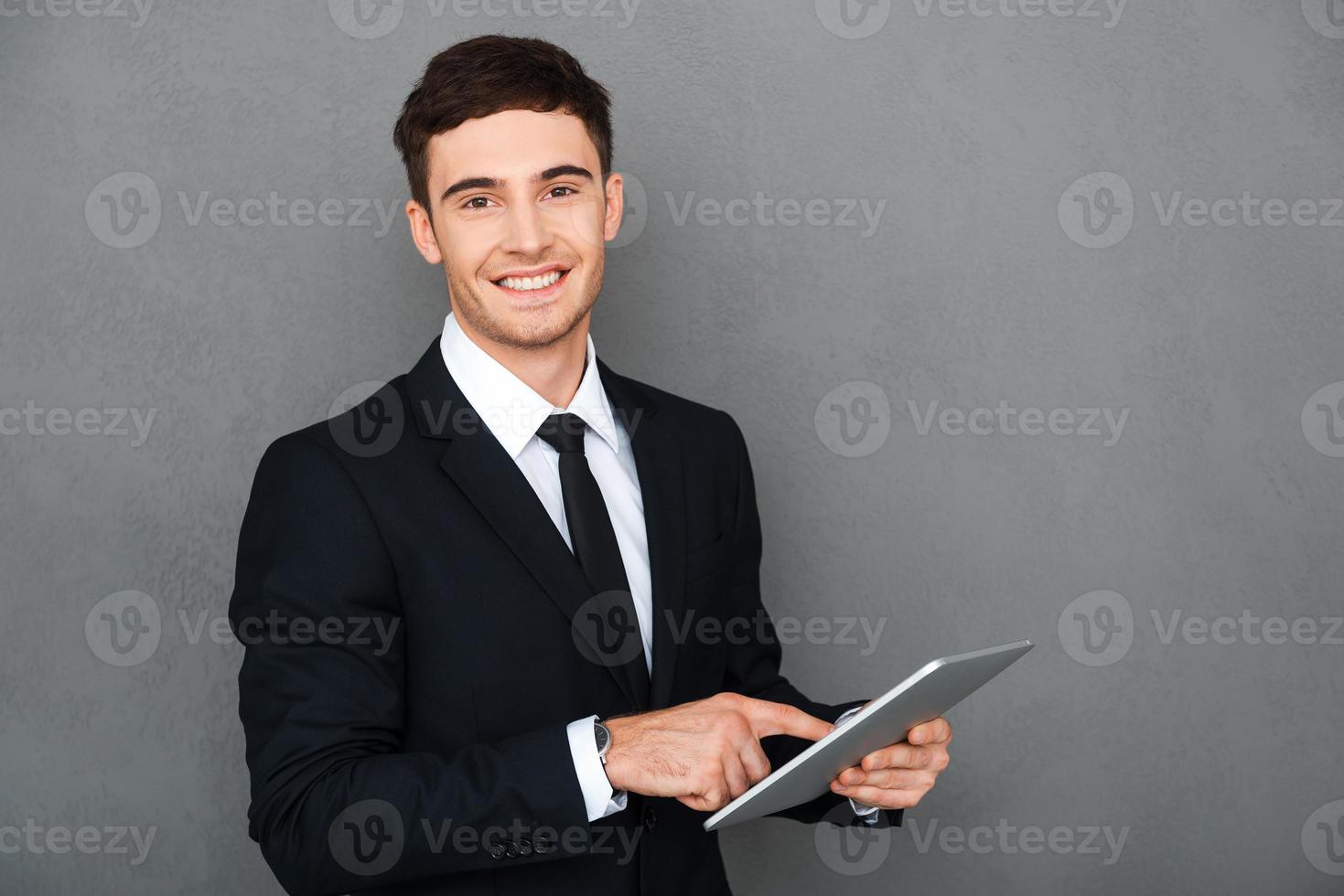 Businessman with digital tablet. Happy young man in formalwear working on digital tablet and looking at camera while standing against grey background photo