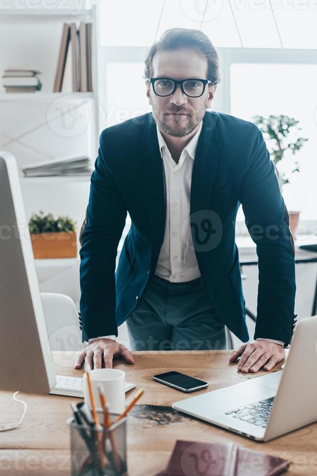 hombre de negocios confiado. apuesto joven con anteojos mirando a la cámara mientras se apoya en la mesa en la oficina creativa foto