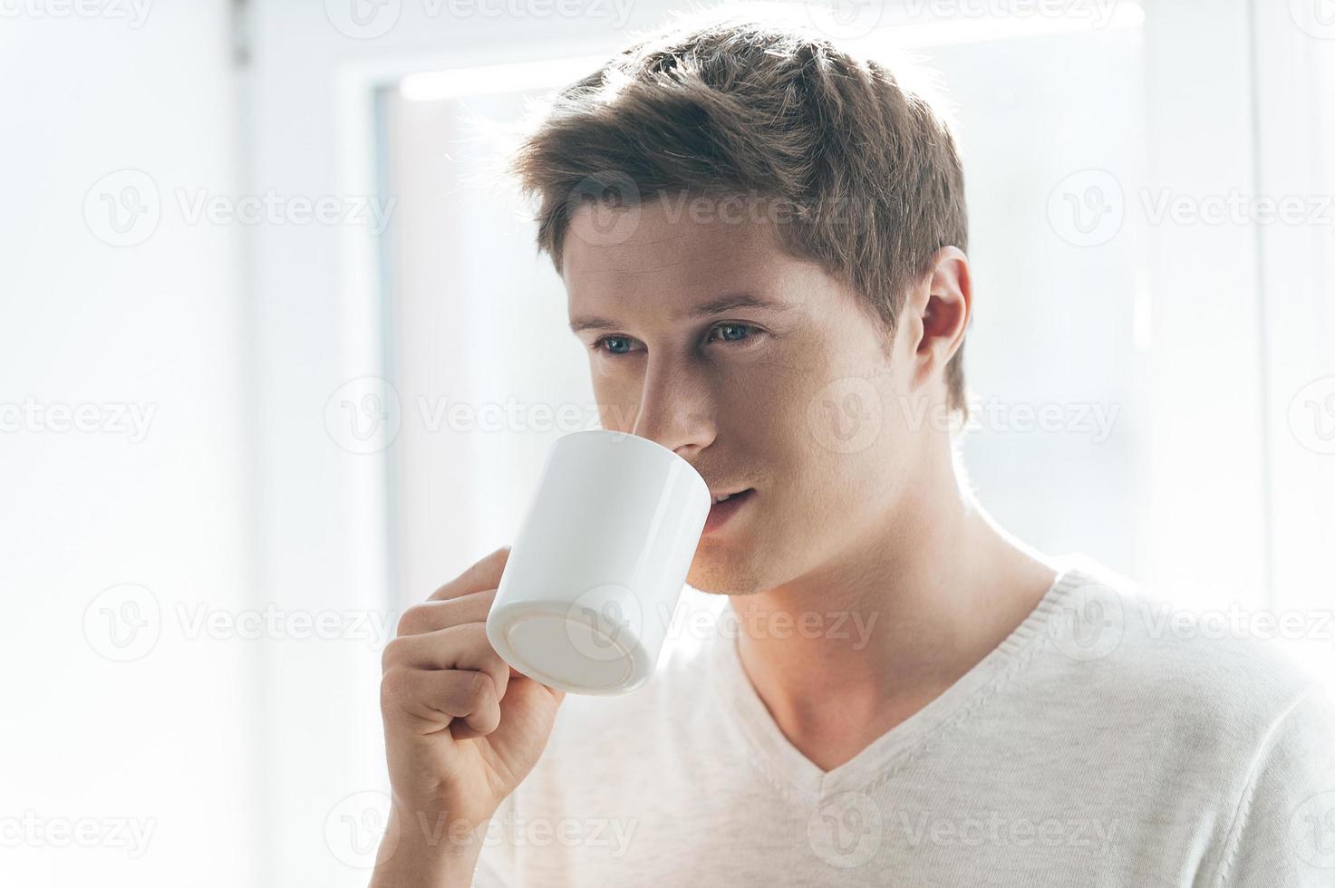 Inspired with cup of fresh coffee. Cheerful young man drinking coffee and looking pensive while standing against window photo
