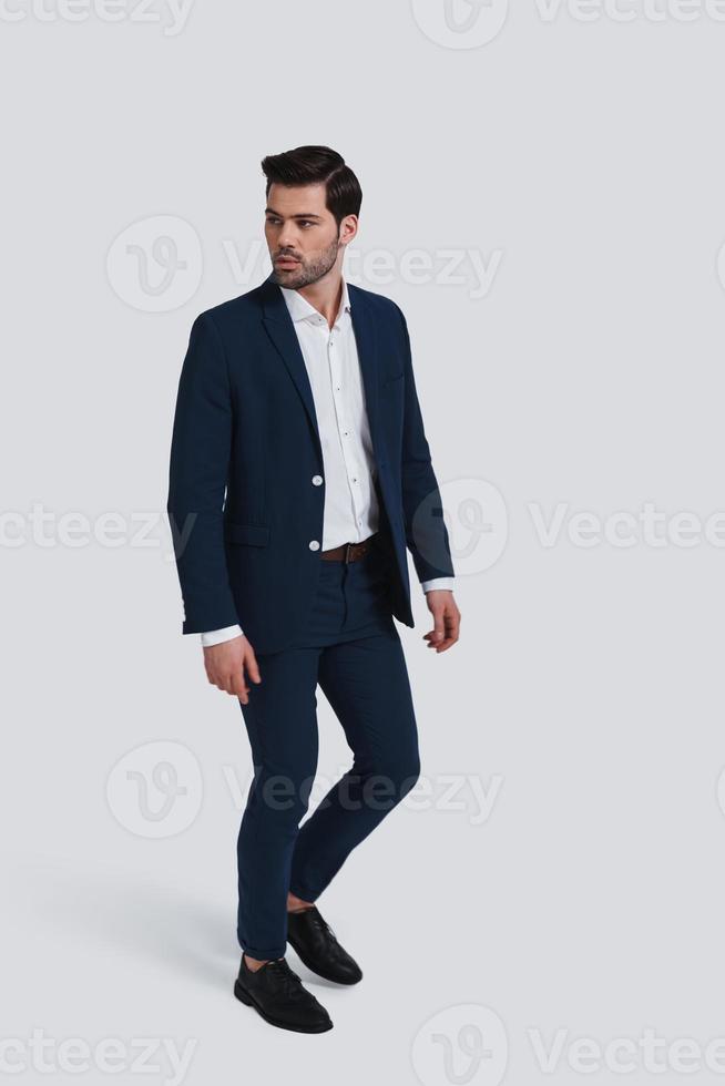 Always perfect. Full length of handsome young man in full suit looking away while walking against grey background photo