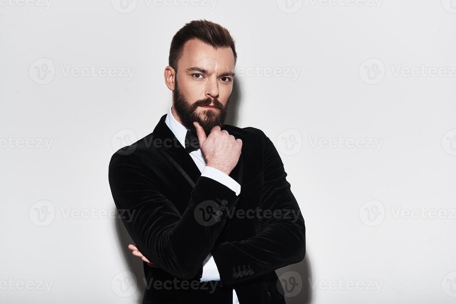 Portrait of confidence. Handsome young man in full suit keeping hand on chin and looking at camera while standing against grey background photo