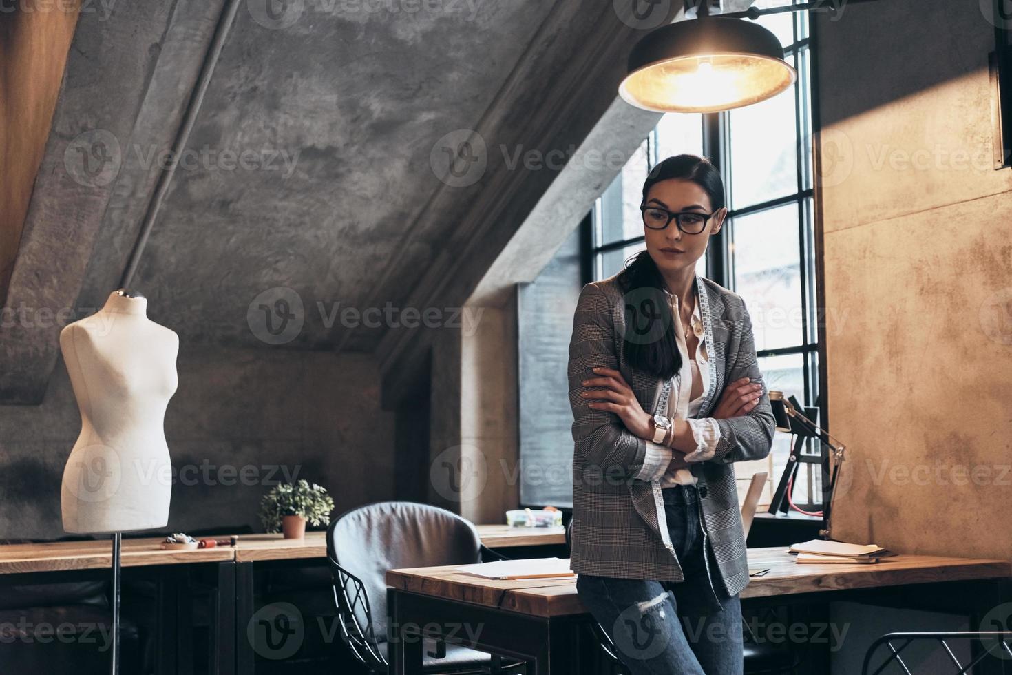 Successful fashion designer. Serious young woman in eyewear looking away and keeping arms crossed while leaning on the desk in her workshop photo