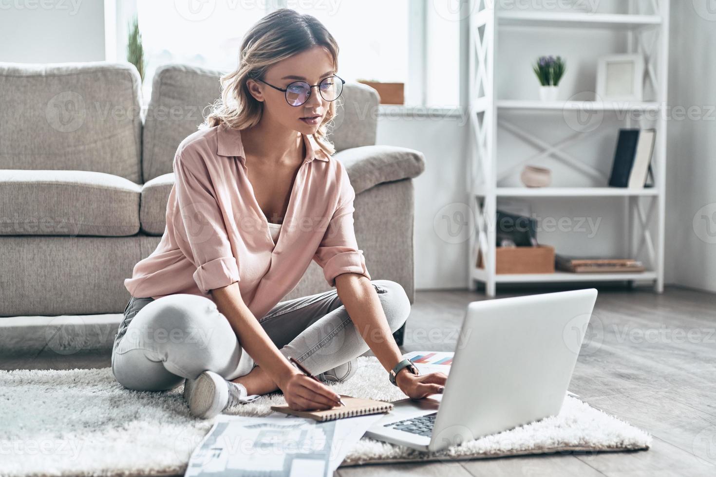 Enjoying her creative job. Thoughtful young woman in eyewear working using computer while flooring at home photo
