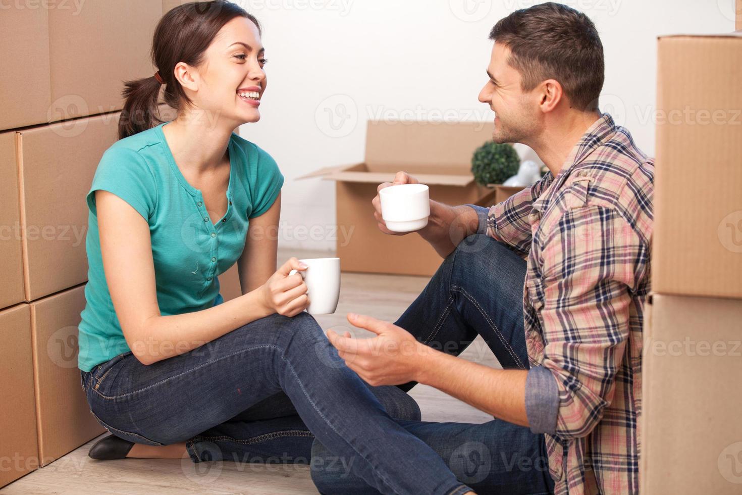 Relaxing in new house. Cheerful young couple sitting on the floor and drinking coffee while cardboard boxes laying all around them photo