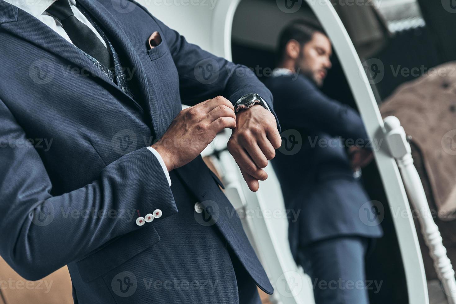 What time is it Close up of young man in full suit checking the time while standing in front of the mirror indoors photo