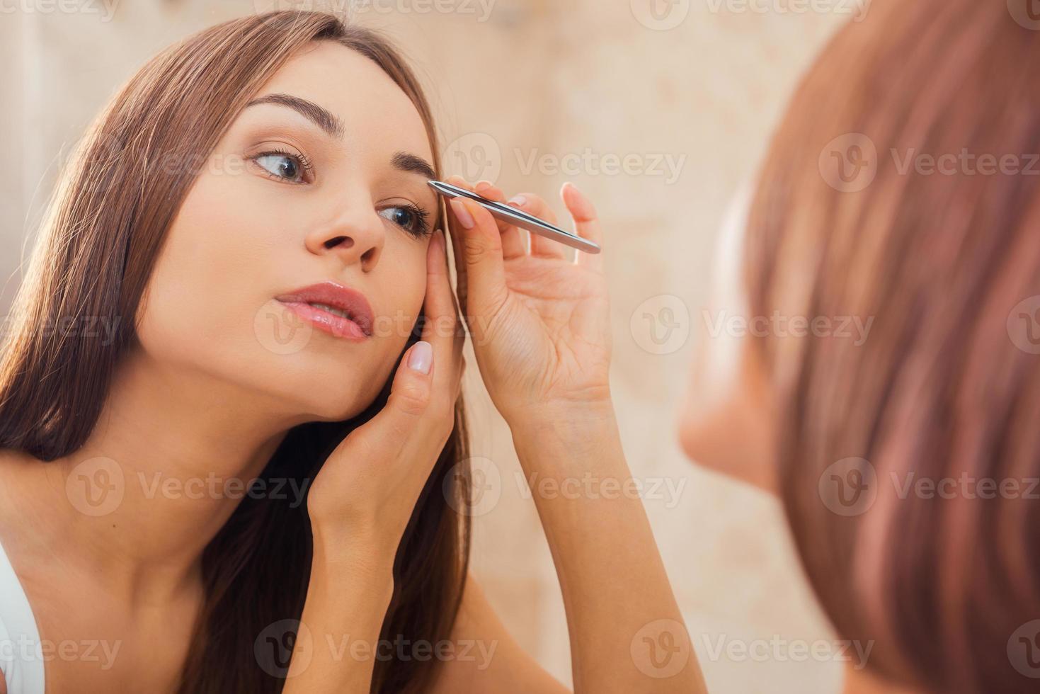 Tweezing eyebrows. Beautiful young woman tweezing her eyebrows while looking at the mirror photo