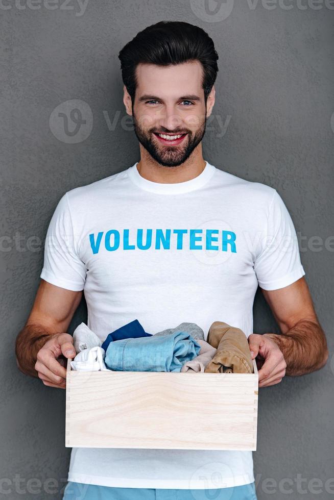 donando con gusto. joven confiado con camiseta de voluntario sosteniendo una caja de donación en sus manos y mirando la cámara con una sonrisa mientras se enfrenta a un fondo gris foto