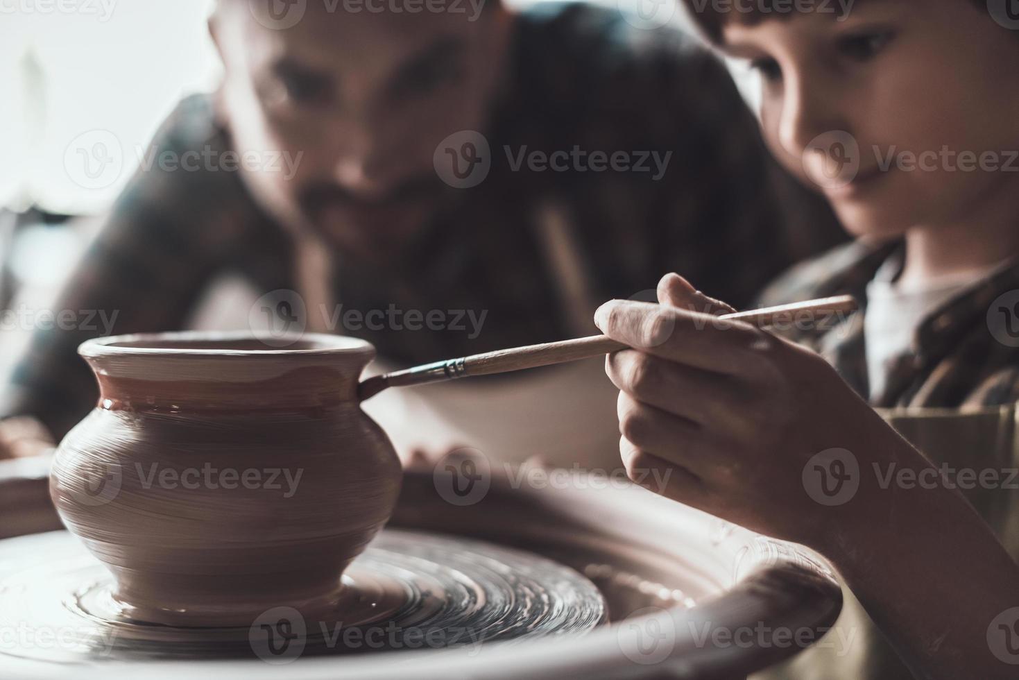 Pottery class. Little boy drawing on ceramic pot at the pottery class while man in apron standing close to him photo