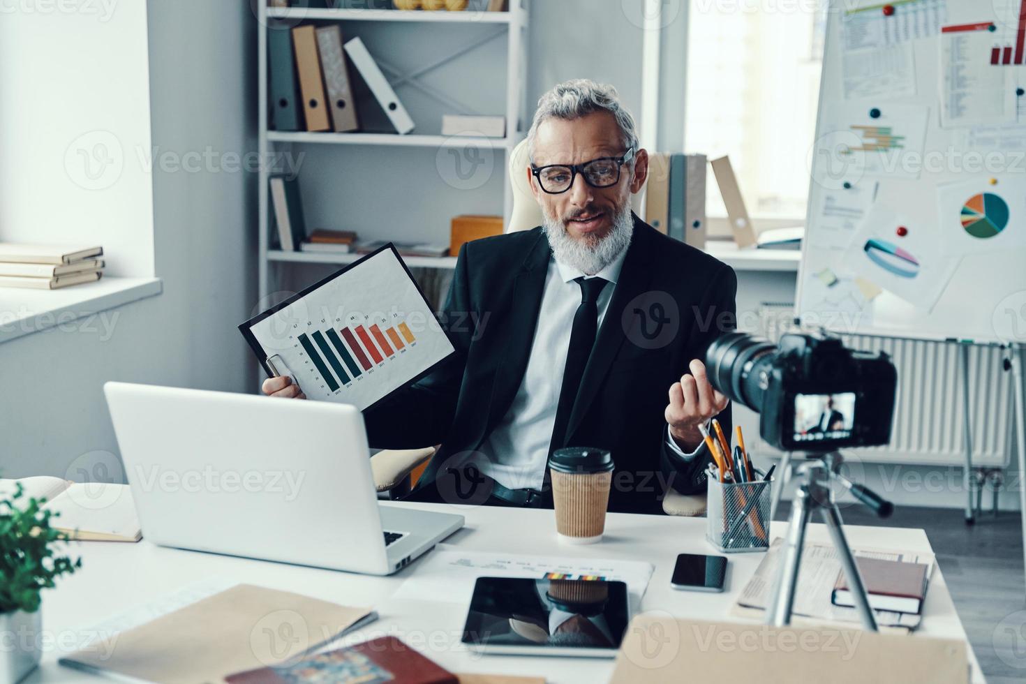 Confident mature man in elegant suit showing chart and sharing business experience while making social media video photo