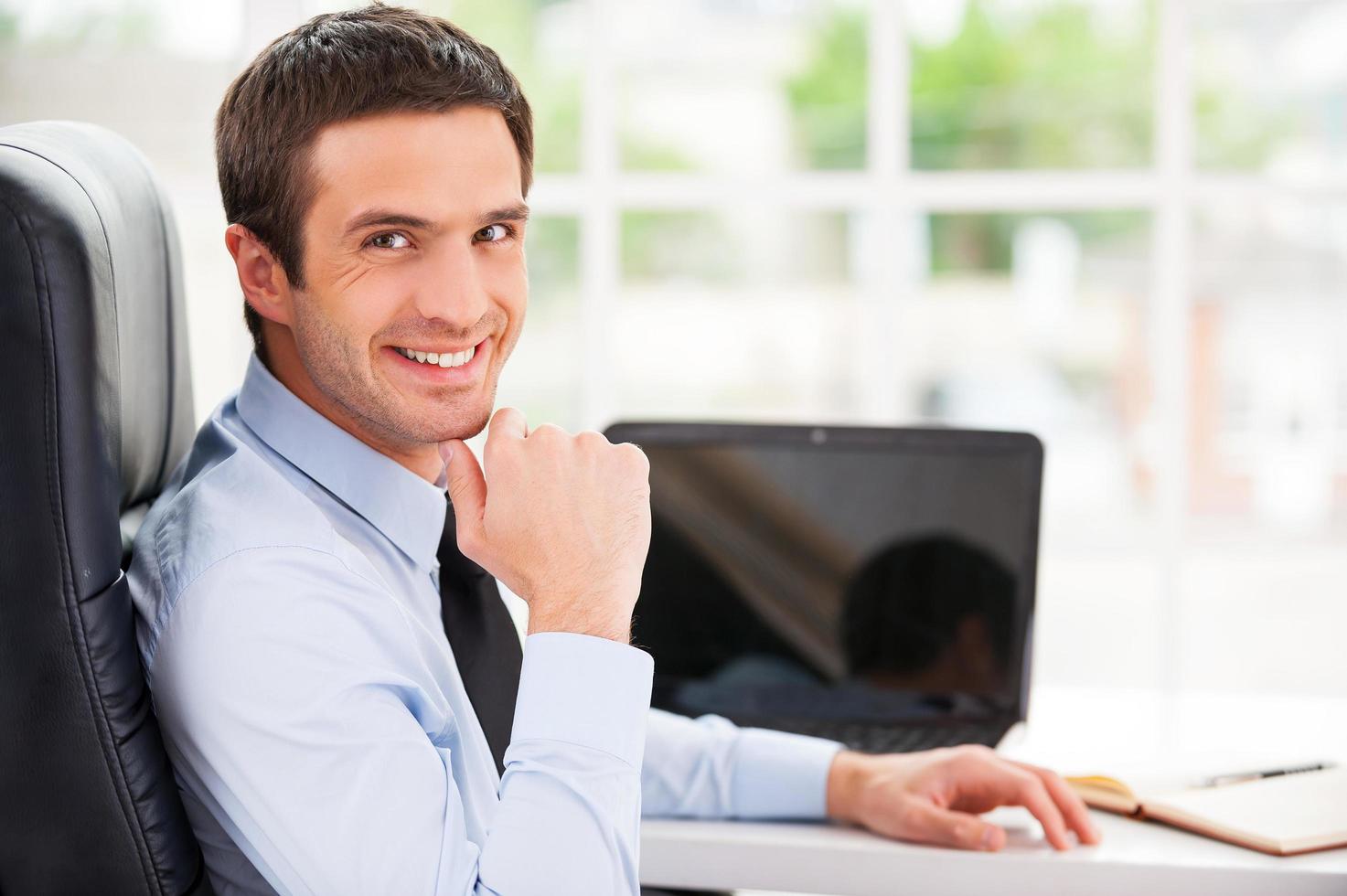 confiado y exitoso. un joven apuesto con ropa formal mirando por encima del hombro y sonriendo mientras se sienta en su lugar de trabajo foto