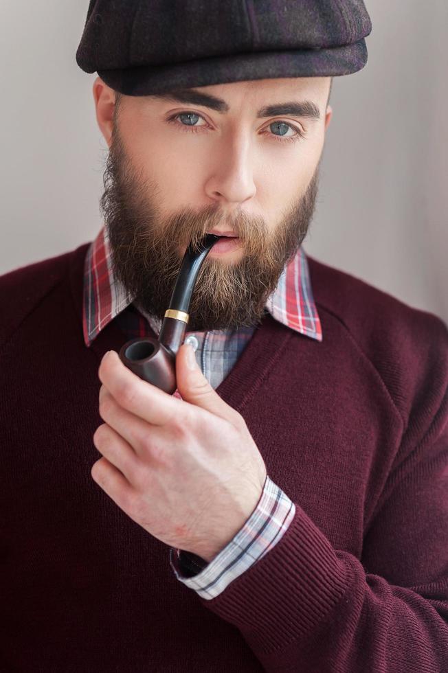 Elegance and masculinity. Portrait of handsome young man in hat smoking a pipe and smiling at camera photo