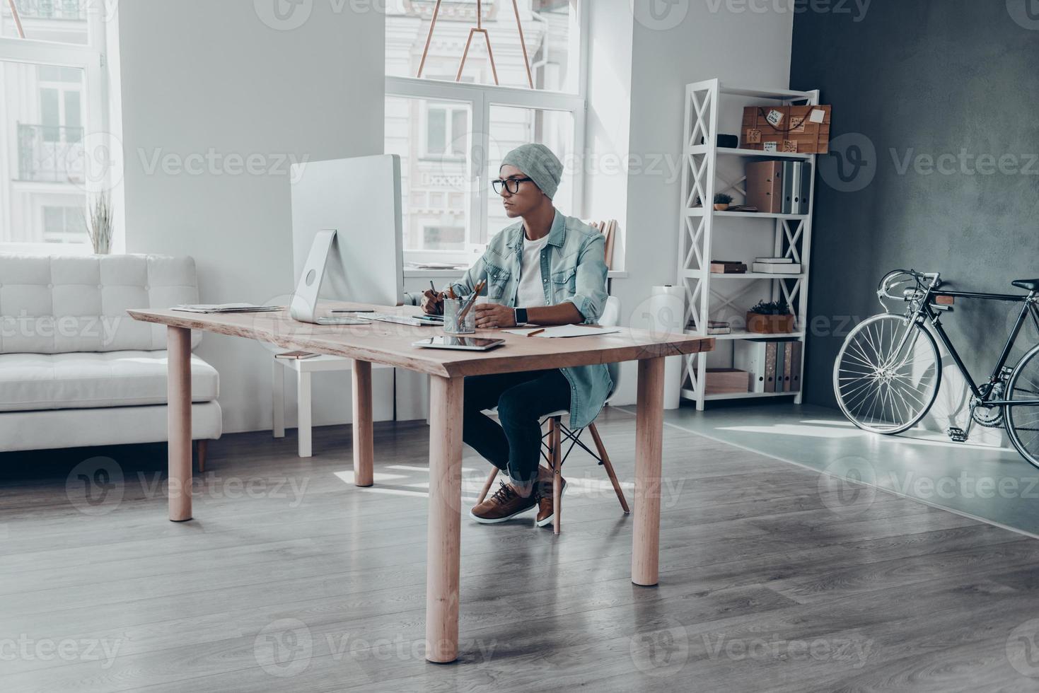 Putting ideas into something real. Handsome young man in eyewear looking at monitor while sitting at his working place in creative office photo