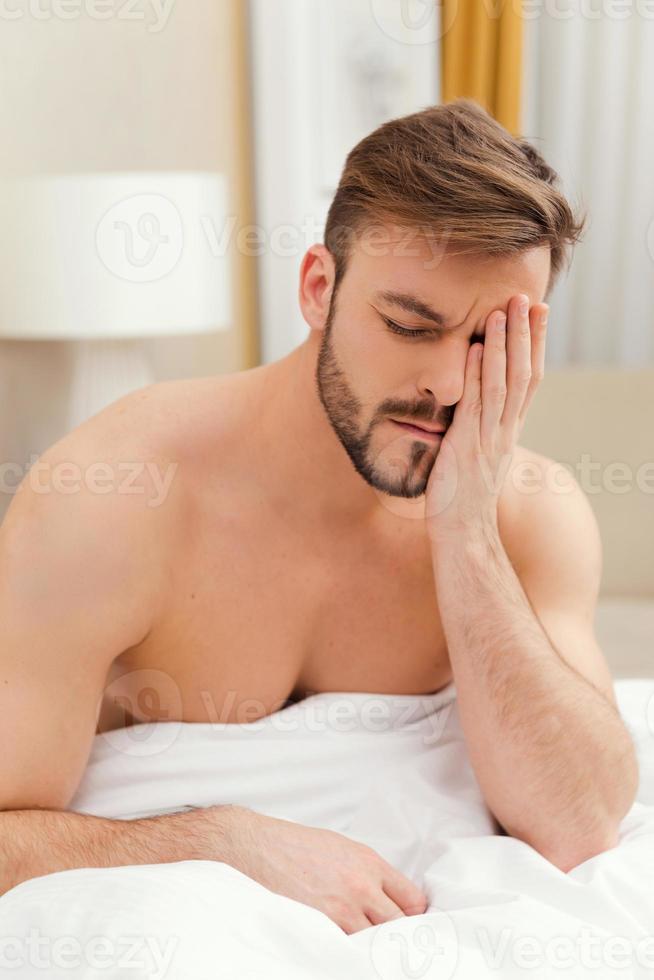 Early wake up. Depressed young man sitting in bed and touching face with hand photo