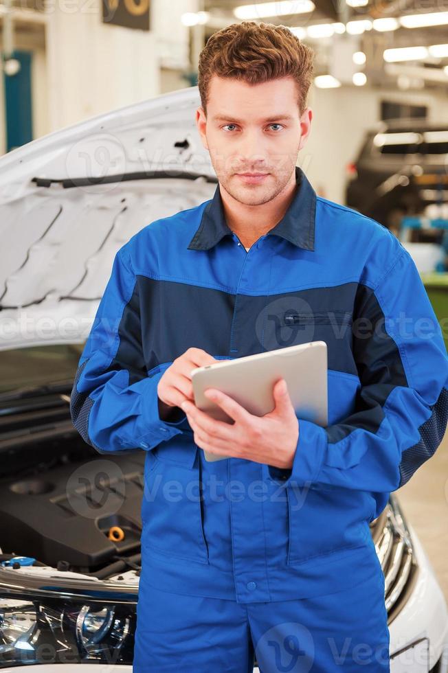 concentrado en su problema. joven seguro de sí mismo trabajando en una tableta digital y mirando la cámara mientras está de pie en el taller con el coche en el fondo foto