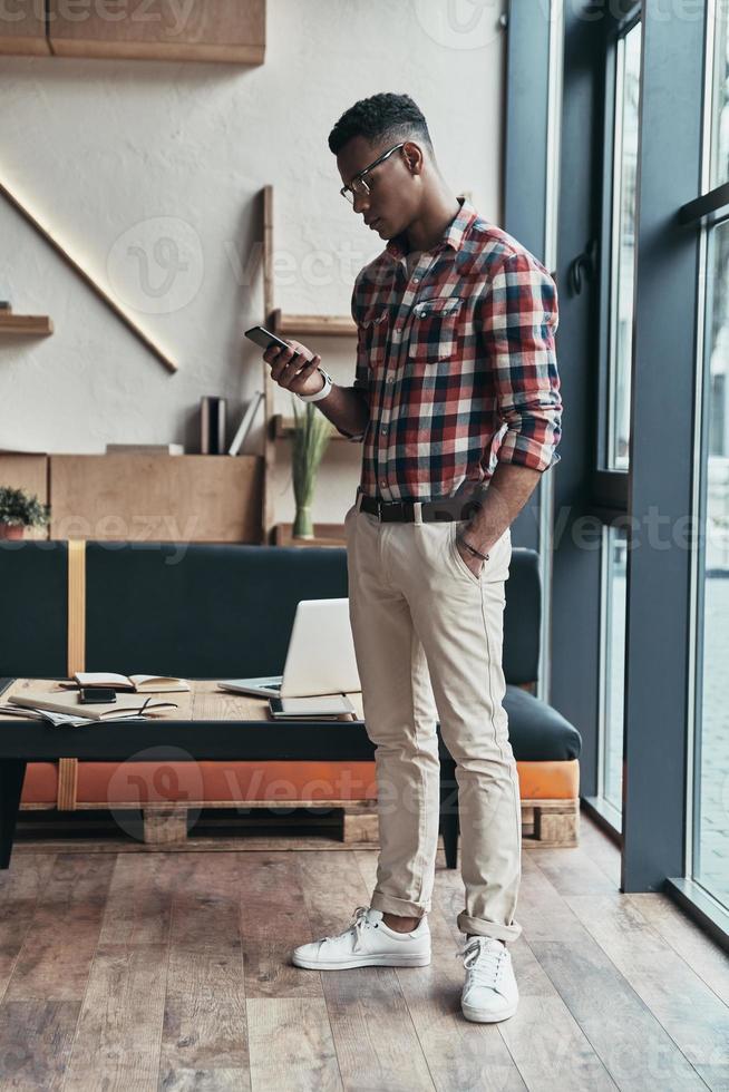 Another business message. Full length of thoughtful young man using his smart phone and keeping hand in pocket while standing in the creative working space photo
