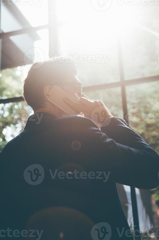 Talking about business. Low angle rear view of confident young man talking on the mobile phone and looking away while standing in front of the big window indoors photo