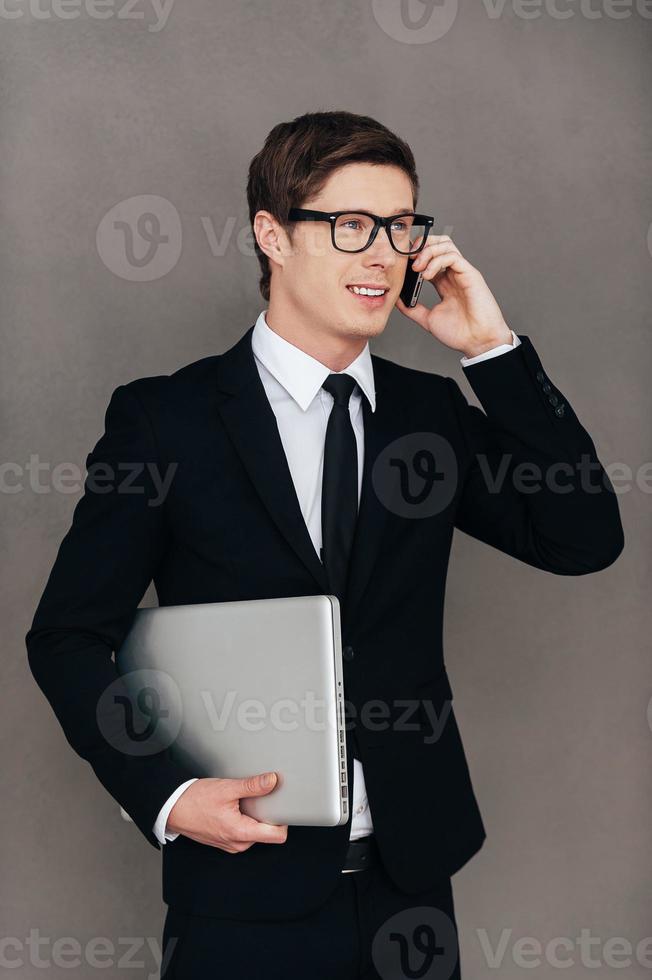 Staying in touch with the colleagues. Confident young man in formalwear carrying laptop and talking on the mobile phone while standing against grey background photo