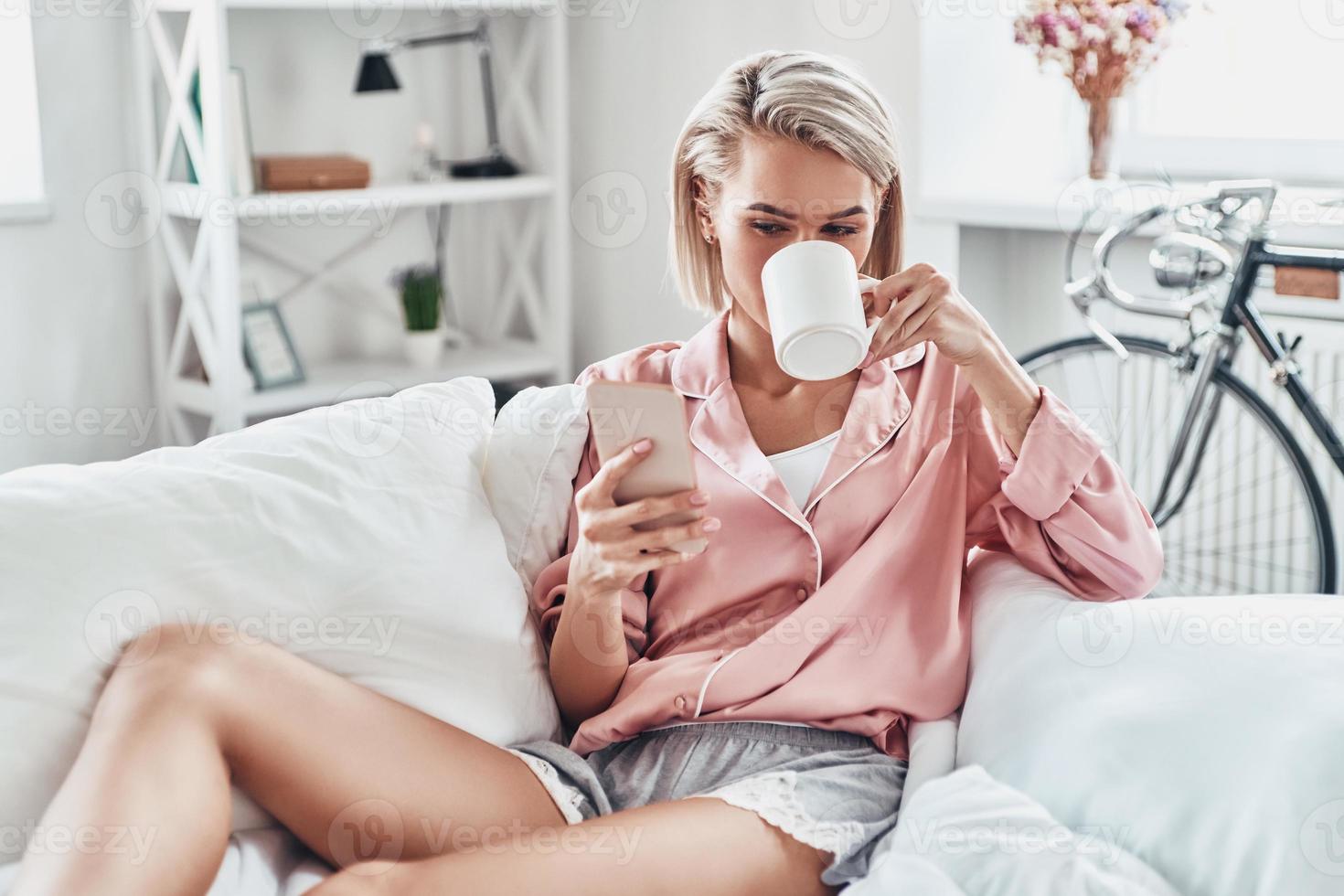 Lazy Sunday. Attractive young woman using smart phone while sitting on bed at home photo