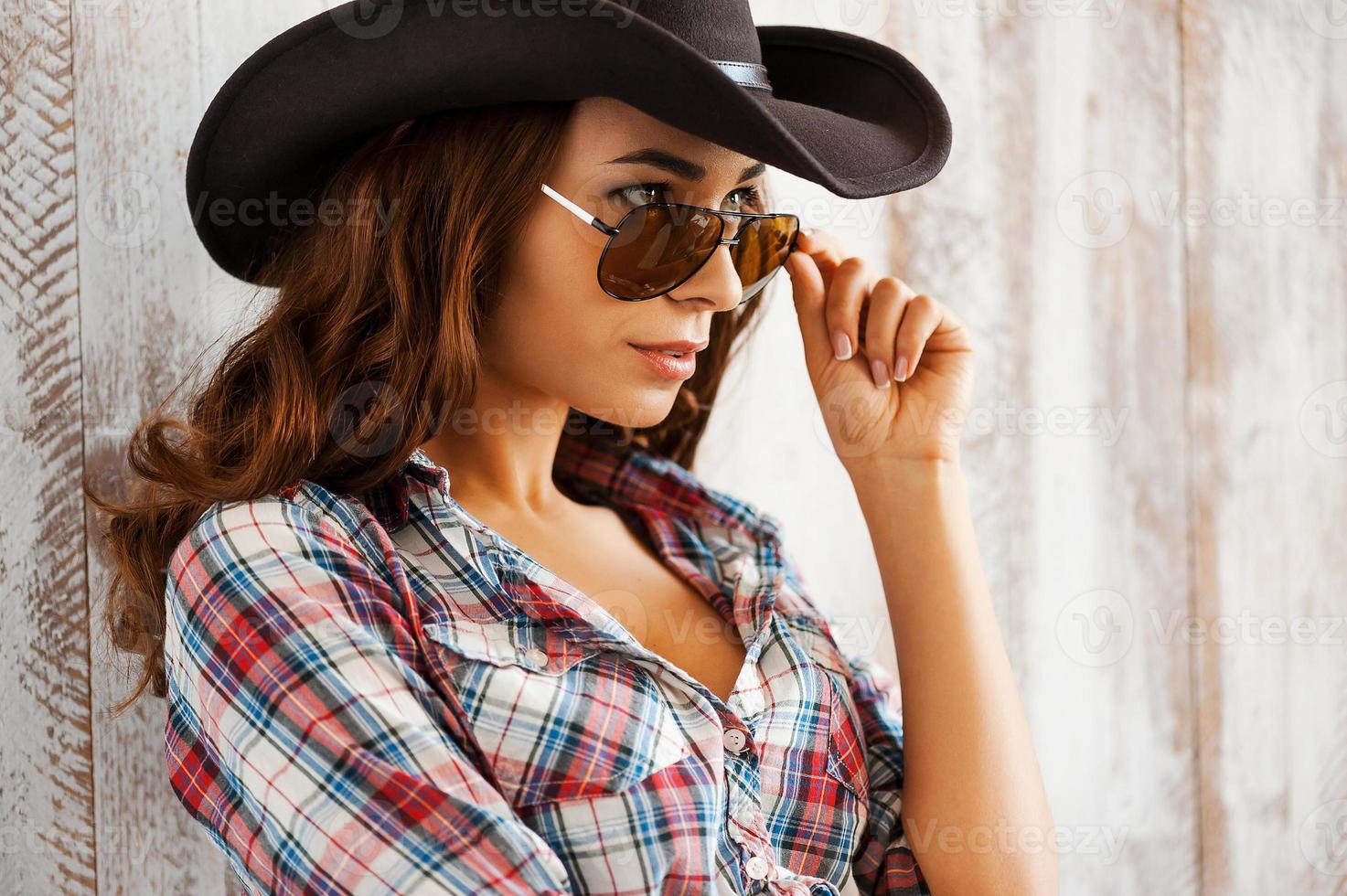 calentando el salvaje oeste. hermosa joven vaquera ajustando sus gafas y mirando a la cámara mientras está de pie contra el fondo de madera foto