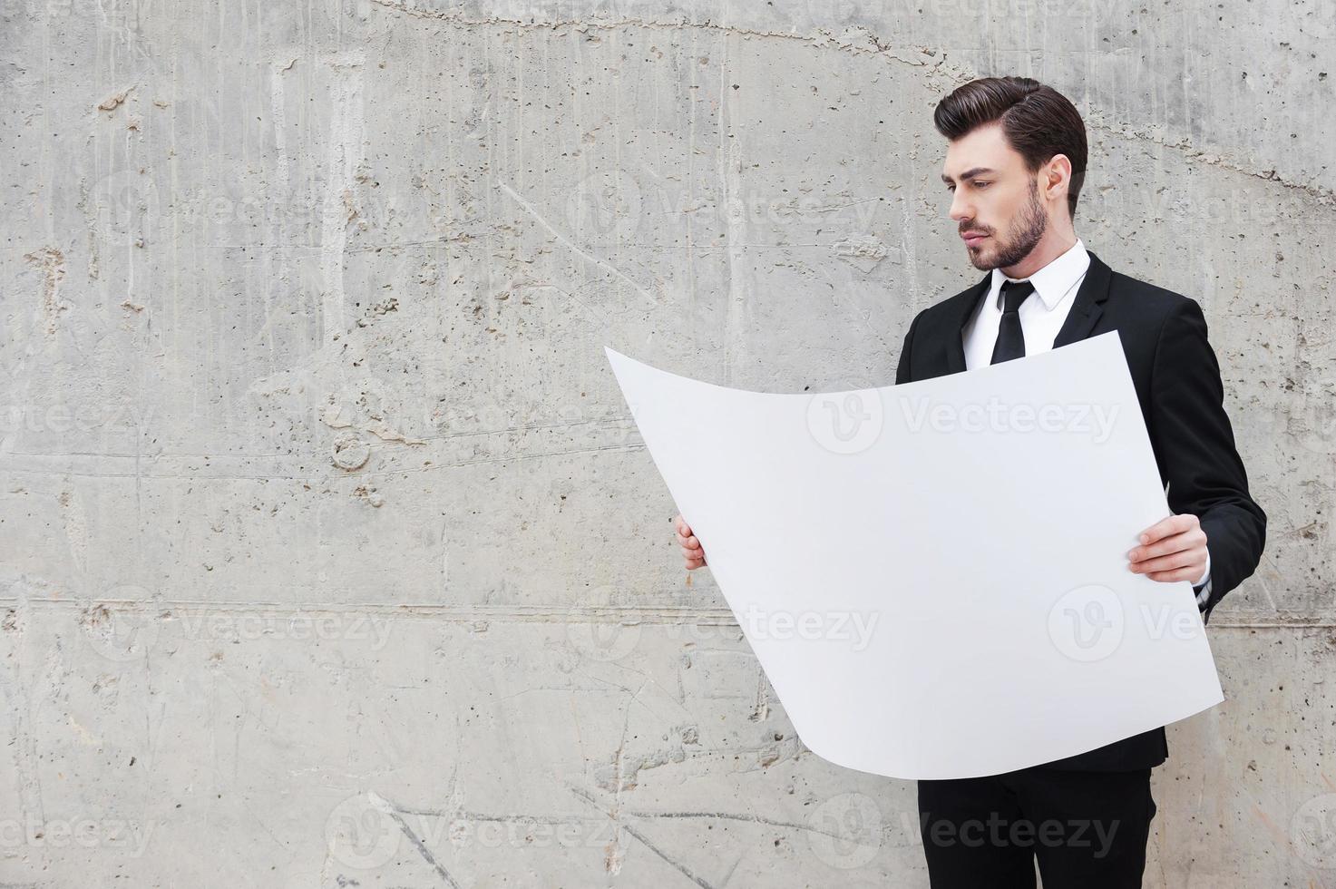 Blueprint for new project. Handsome young man examining blueprint while standing outdoors and against the concrete wall photo