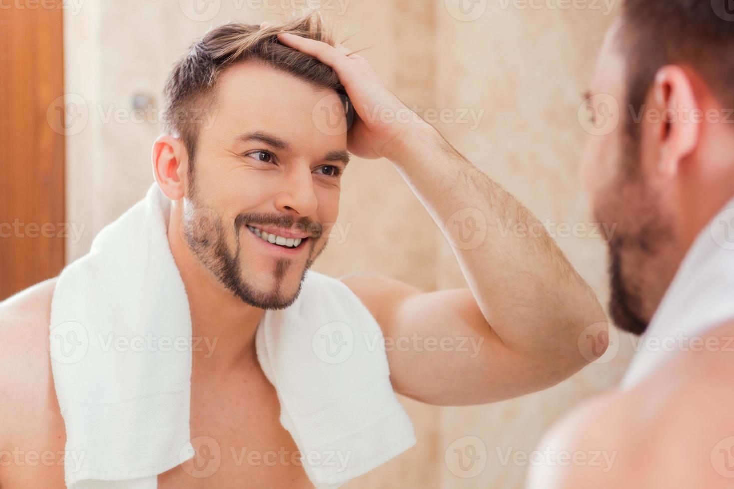 Good morning to me. Handsome young man touching his hair with hand and smiling while standing in front of the mirror photo