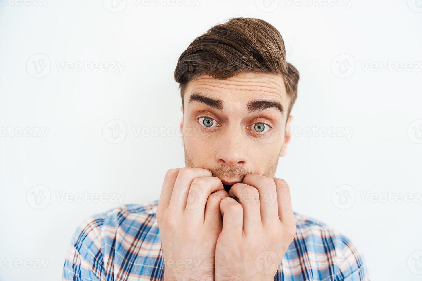 Feeling so scared. Nervous young man biting nails and looking at camera while standing against white background photo