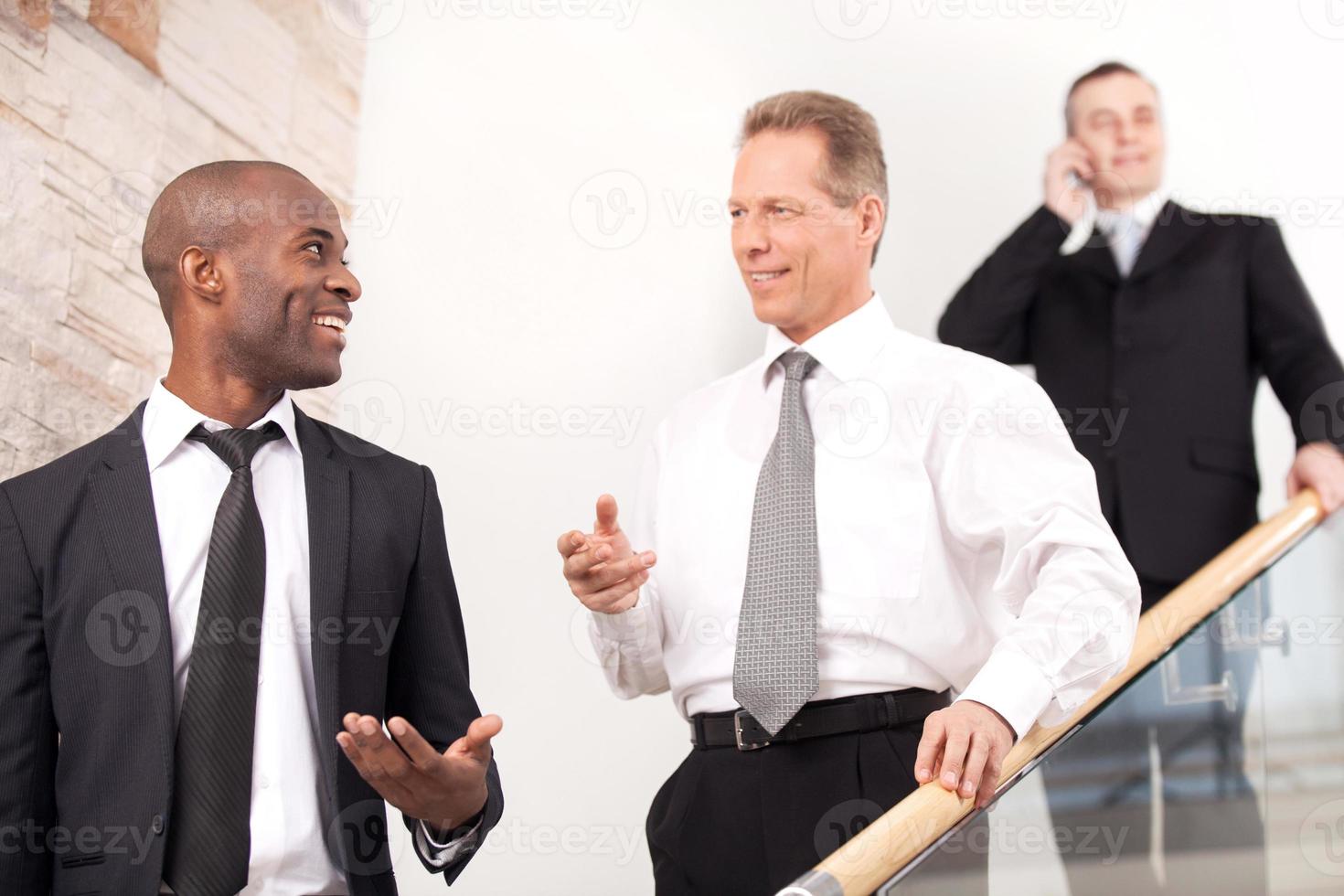 Business people on staircase. Three cheerful man in formalwear moving downstairs while two of them talking to each other and smiling photo