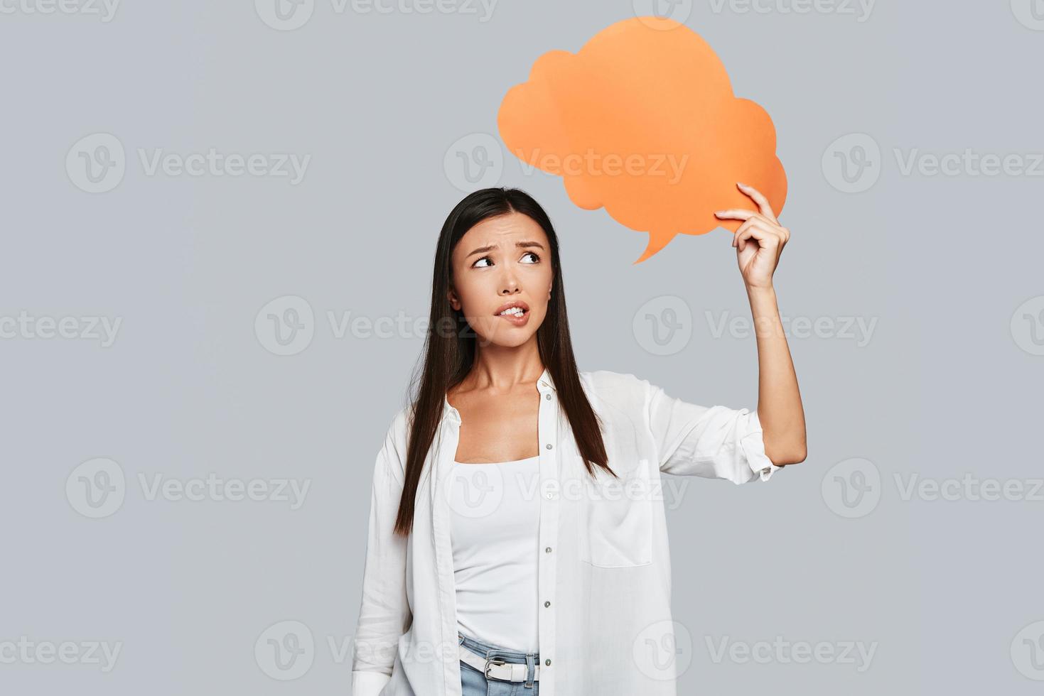 Uncertainty. Beautiful young Asian woman biting lip while standing against grey background photo