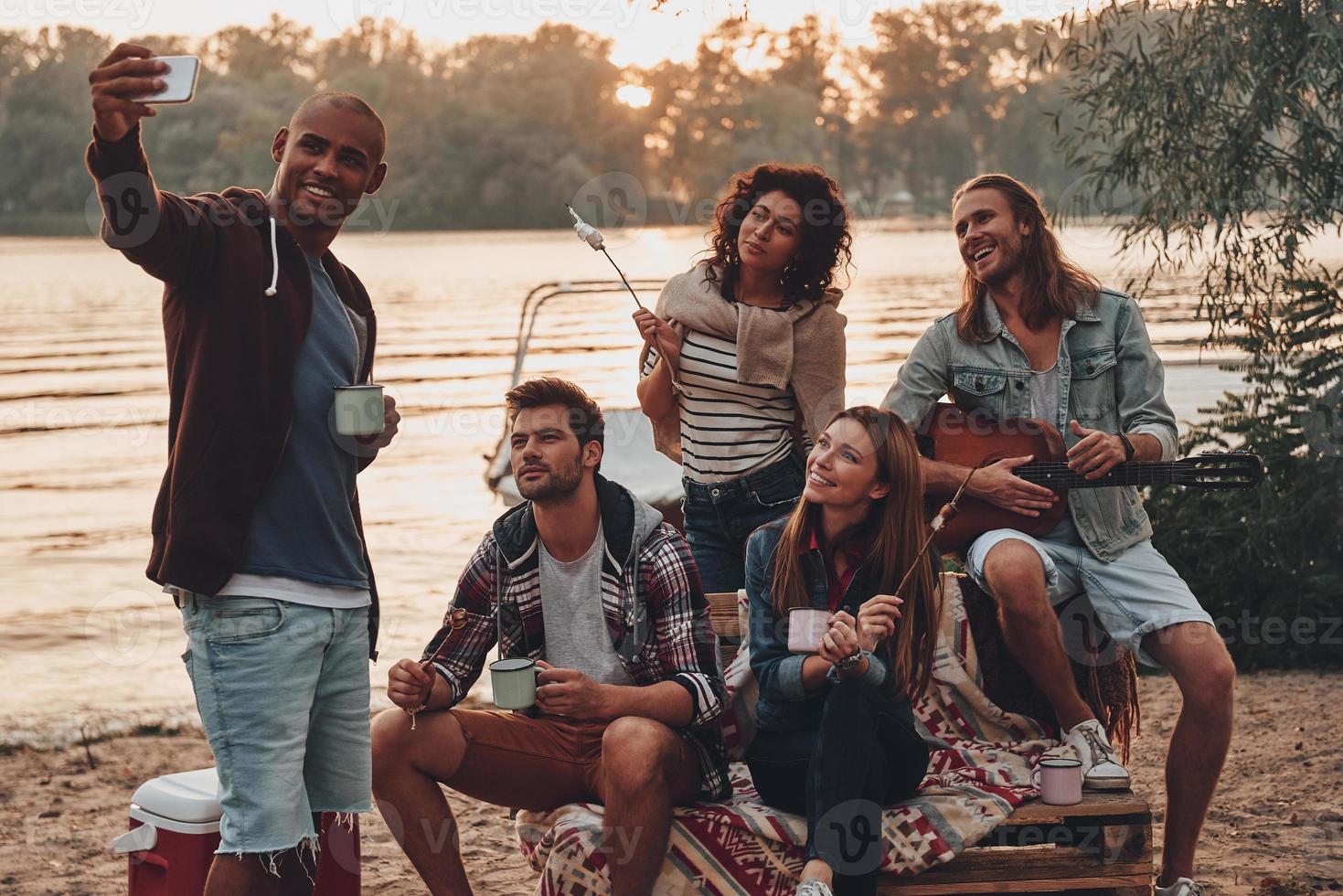 Group of young people in casual wear smiling and taking selfie while enjoying beach party near the lake photo