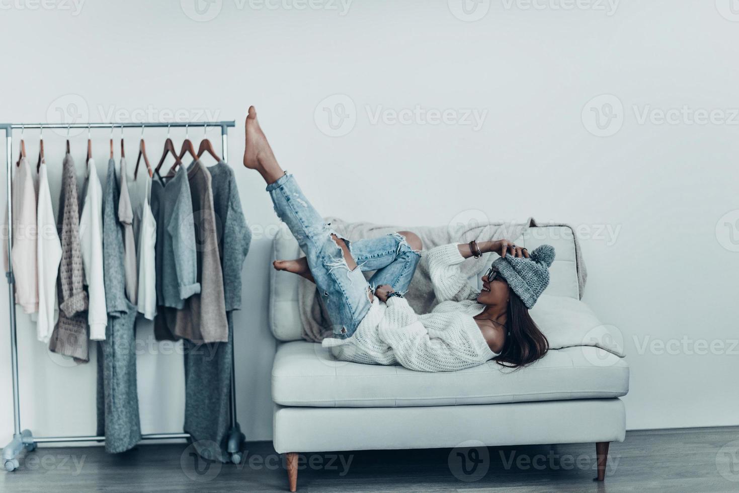 Having fun at home.  Playful young woman in casual wear and knit hat touching her head with hand and putting feet up while lying on sofa at home near her clothes hanging on the racks photo