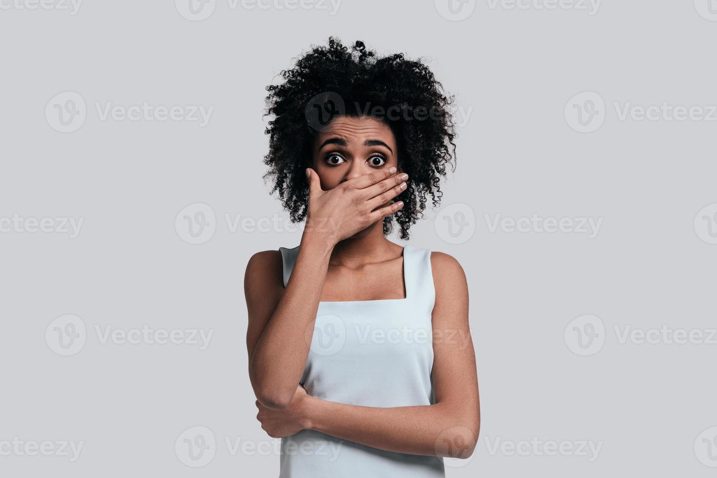 Surprised young African woman in casual wear covering mouth with hand and looking at camera while standing against grey background photo