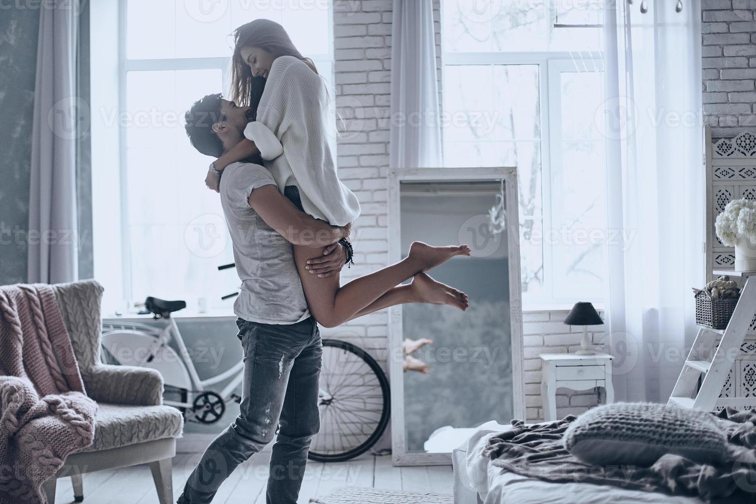Expressing their love. Handsome man in white T-shirt carrying young attractive woman while spending free time at home photo