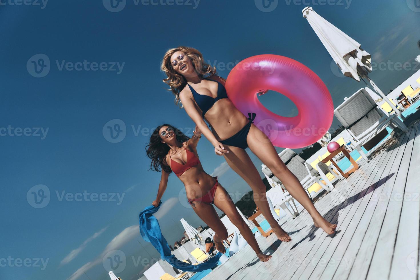 Moments of happiness. Full length of two beautiful young women in bikini running near swimming pool together while one of them carrying inflatable ring photo