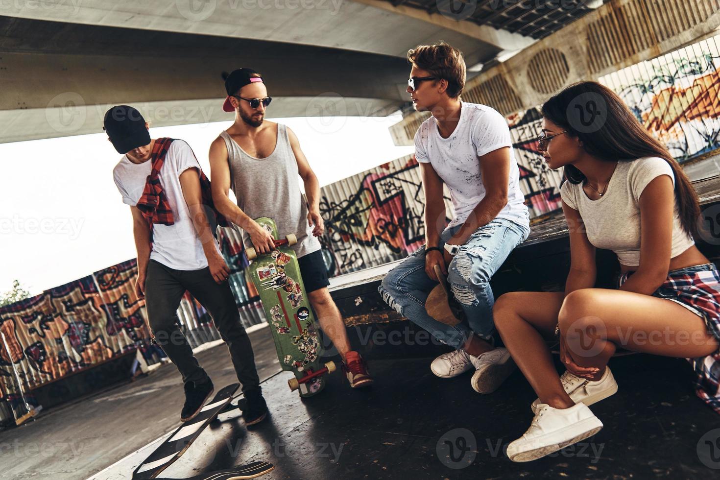 Taking a break. Group of young modern people talking while spending time at the skateboard park outdoors photo