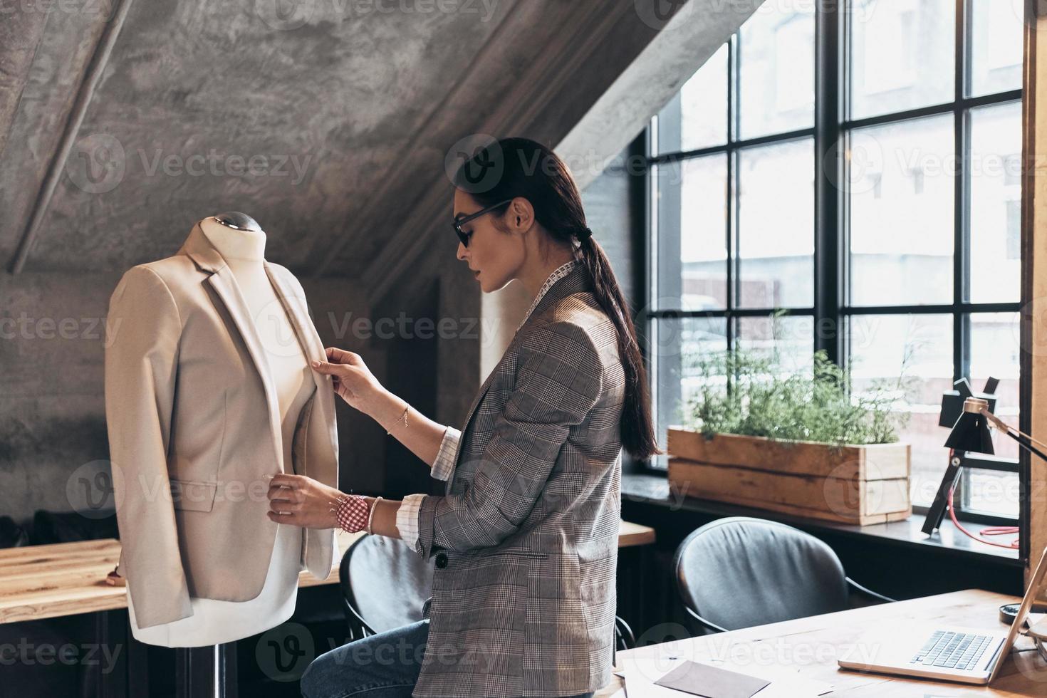 Ready to make your dream come true. Serious young woman in eyewear adjusting a jacket on mannequin while standing in her workshop photo