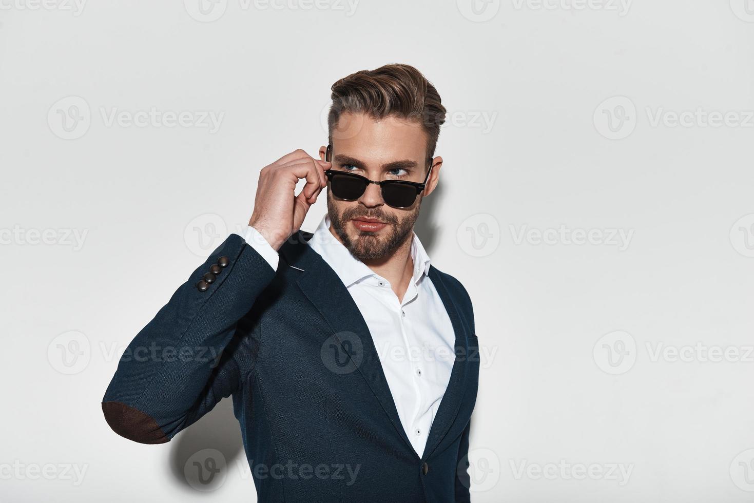 sintiéndose confiado en su estilo. un joven apuesto con ropa formal ajustando sus gafas y mirando hacia otro lado mientras se enfrenta a un fondo gris foto